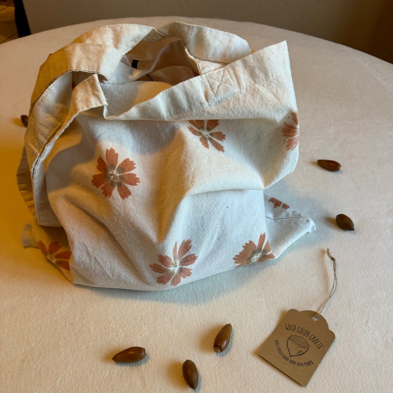 May include: A cream-colored tote bag with a floral pattern in shades of orange and brown. The bag is open, revealing the interior. Several acorns are scattered around the bag. A tag reads "Wild Color Crafts" with a logo of an acorn.