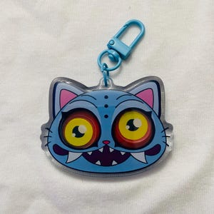 Peut inclure: Un porte-clés bleu en forme de chat avec un design de dessin animé. Le chat a de grands yeux jaunes avec des pupilles rouges, des dents pointues et une bouche violette. Le porte-clés a un fermoir bleu.