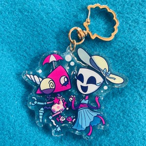 May include: A clear acrylic keychain featuring a colorful illustration of two cartoon characters. One character is pink and triangular with a small umbrella, while the other is white and blue wearing a sun hat. It has a gold-toned shell-shaped clasp.