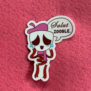 May include: A white sticker featuring a cartoon skeleton character wearing a pink beret and scarf. The character has large, expressive eyes with tears and a speech bubble that says "Salut ZOOBLE."
