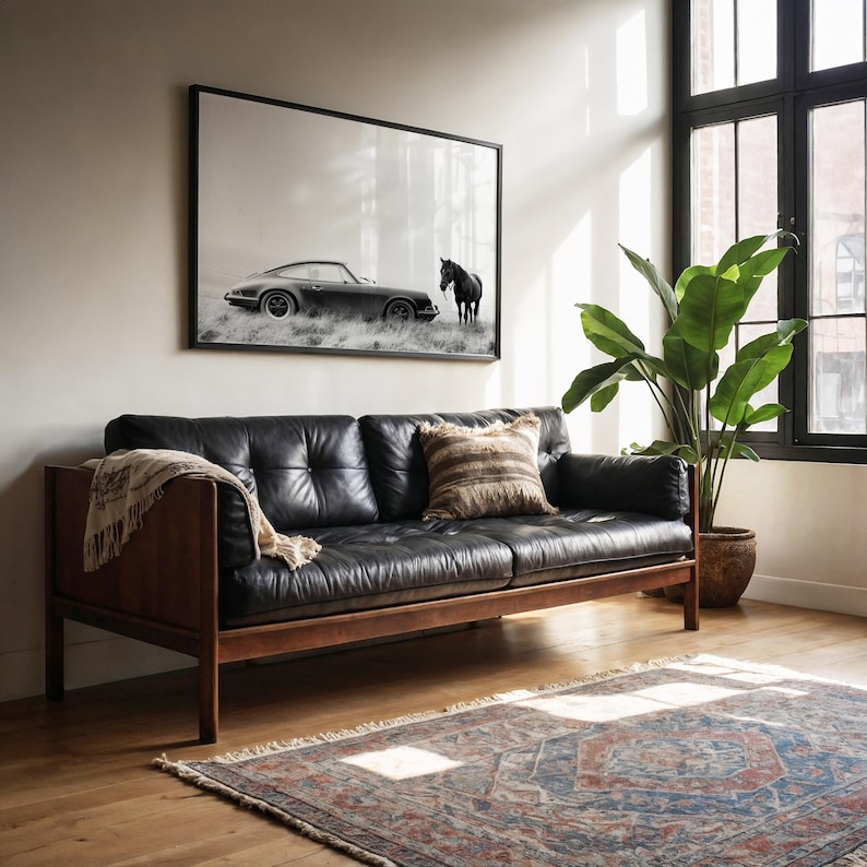 K&ouml;nnte beinhalten: Ein schwarzes Ledersofa mit Holzarmlehnen und -beinen steht in einem Wohnzimmer mit einem gro&szlig;en Fenster und einem Teppich. Ein Schwarzwei&szlig;foto eines Autos und eines Pferdes h&auml;ngt an der Wand &uuml;ber dem Sofa.