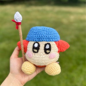 May include: A handmade crochet plush toy of a character with a blue hat, red arms, and pink cheeks. The character is holding a spear with a white tip and red ribbon. The toy is made of beige, yellow, red, and blue yarn.