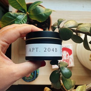 May include: A black candle tin with a gold rim and a white label that reads "APT. 204B". The tin is held in a person's hand, with green plants in the background.