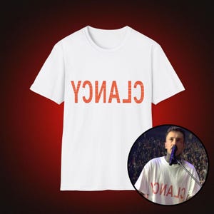 May include: A white short-sleeved t-shirt featuring the word CLANCY printed in large, orange, distressed-style capital letters. The text is oriented upside down. A circular inset shows a person wearing the same white t-shirt on a stage.