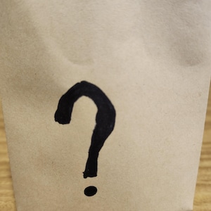 May include: A light brown paper bag featuring a single, hand-drawn black question mark symbol centered on the front surface. The bag is made of a matte, textured paper material and displays a simple, bold graphic design in black ink.