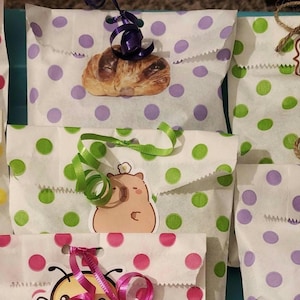 May include: A collection of seven white paper gift bags featuring colorful polka dots in pink, yellow, purple, and green. Each bag is decorated with a unique sticker, including a coffee cup, a pastry, a dragon, a bear, and a rooster, with ribbons.