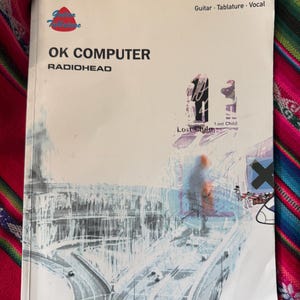 May include: A music book titled "OK Computer" by Radiohead. The cover features a grayscale illustration of a highway and the text "Guitar, Tablature, Vocal". The book also includes the song title "Lost Child".