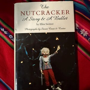 May include: A book cover for "The Nutcracker: A Story & A Ballet" by Ellen Switzer. The cover features a costumed figure from the ballet, set against a dark background with scattered lights. The figure wears a mask and a military-style jacket.