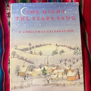 May include: A hardcover book titled "The Night the Stars Sang: A Christmas Celebration." The cover features a winter village scene with snow-covered houses, trees, and a church under a starry sky. The book is set against a colorful patterned background.