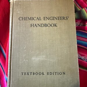 May include: A vintage textbook titled "Chemical Engineers' Handbook" in a light olive green cover. The words "Textbook Edition" are printed at the bottom. The book is set against a colorful, patterned textile background.