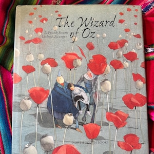 May include: A hardcover book titled "The Wizard of Oz" with illustrations of red poppies and characters. The cover art features a light blue background with the book title in elegant script. Published by North-South Books.