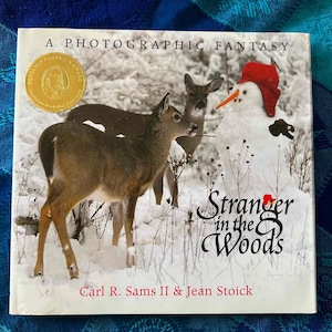 May include: A book cover with a snowy winter scene featuring a snowman wearing a red hat and two deer. The title of the book is "Stranger in the Woods" by Carl R. Sams II & Jean Stoick.