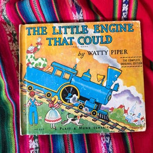 May include: Vintage children's book titled "The Little Engine That Could" by Watty Piper. The cover features a blue train illustration with a clown, children, and a dog. The book is a Platt & Munk Classic.