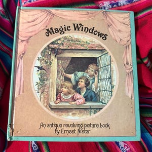 May include: Antique children's book titled "Magic Windows" by Ernest Nister. The cover features a circular illustration of children looking out a window. The book has a light blue border and a pink and beige cover.