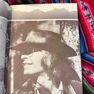 May include: A sepia-toned photograph of a person wearing a hat and sunglasses, smiling. The person is wearing a jacket and a collared shirt. The background features a mountain range silhouette.