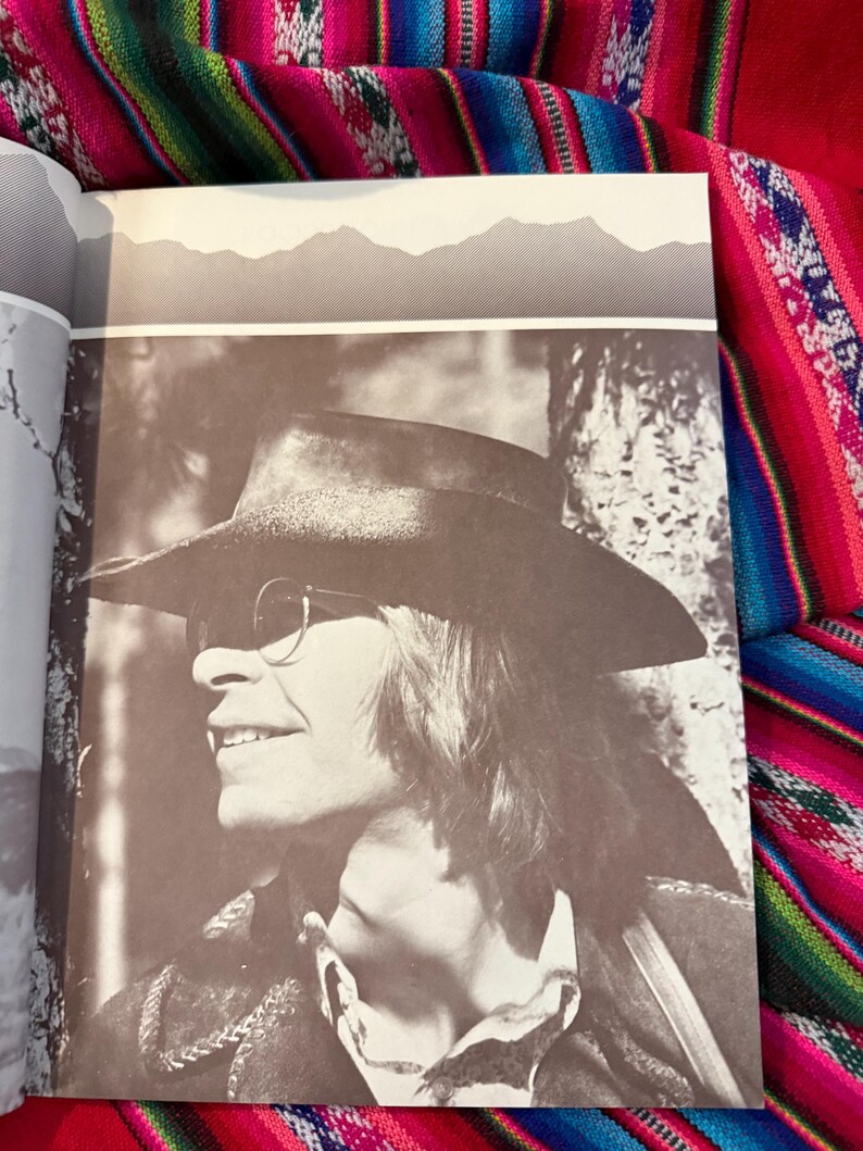 May include: A sepia-toned photograph of a person wearing a hat and sunglasses, smiling. The person is wearing a jacket and a collared shirt. The background features a mountain range silhouette.