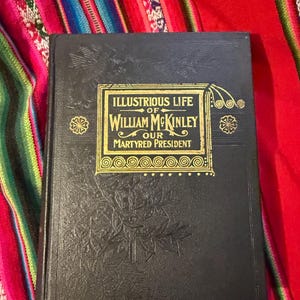 May include: A vintage, hardcover book titled "Illustrious Life of William McKinley Our Martyred President." The cover is black with gold lettering and decorative elements. The book is set against a colorful, patterned textile background.