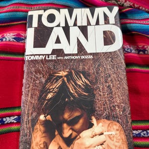 May include: A book titled "TOMMY LAND" by Tommy Lee with Anthony Bozza. The cover features a sepia-toned image of a person with tattoos, holding a cigarette. The book is set against a colorful, patterned textile.
