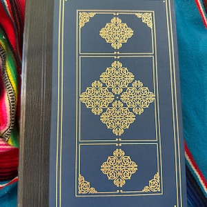May include: A hardcover book with a dark blue cover and gold decorative accents. The cover features three rectangular panels with ornate gold designs. The spine is black, and the book is resting on a colorful patterned textile.