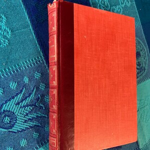 May include: A vintage book with a red cover and spine. The spine is a darker shade of red with gold lettering and decorative elements. The book's cover is a textured, solid red. The book is resting on a blue and teal patterned surface.