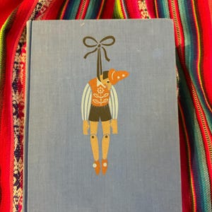 May include: A vintage book with a light blue cover featuring a whimsical illustration of a marionette doll. The doll has an orange hat, vest, and brown legs. The marionette is suspended by a black bow at the top.