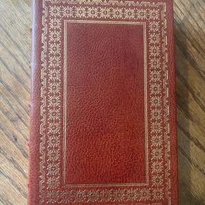 May include: A brown leather-bound book with a gold-coloured decorative border around the cover.