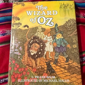 May include: A hardcover copy of "The Wizard of Oz" children's book. The cover features illustrations of the Lion, Scarecrow, Dorothy, and the Tin Man on a yellow and green background. The title is in large, bold letters.