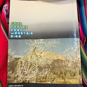 May include: A vintage book cover featuring the text "John Denver Rocky Mountain High" in green and blue. The lower half displays a scenic mountain landscape with snow-covered trees. The Cherry Lane Music Co. logo is visible.