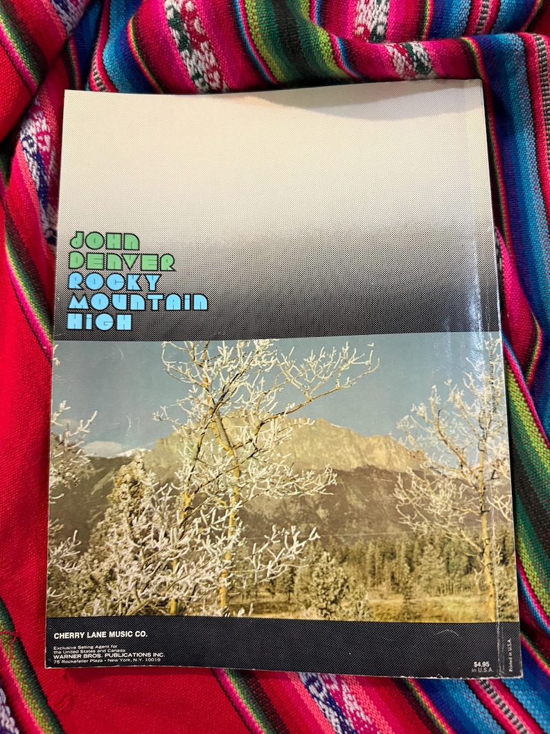 May include: A vintage book cover featuring the text "John Denver Rocky Mountain High" in green and blue. The lower half displays a scenic mountain landscape with snow-covered trees. The Cherry Lane Music Co. logo is visible.