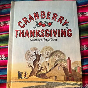 May include: A vintage children's book titled "Cranberry Thanksgiving" by Wende and Harry Devlin. The cover features a colorful illustration of a house, trees, and two children walking in a snowy landscape.
