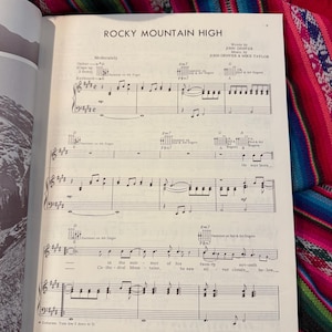 May include: Sheet music for "Rocky Mountain High" by John Denver, featuring musical notation for guitar and keyboard. The page is from a vintage songbook, with the title and composer's names at the top.