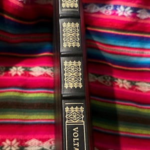 May include: A black book spine with gold lettering and decorative accents. The book title "Candide" is visible at the top, and "Voltaire" at the bottom. The spine has a textured appearance.