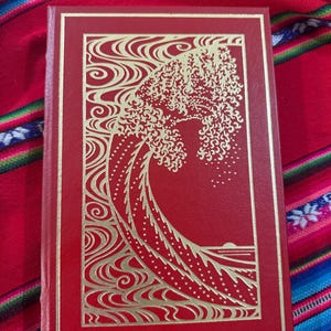 May include: A red book with a gold embossed design of a wave. The cover has a gold border and the background is a colorful textile. The book's design is inspired by Japanese art.