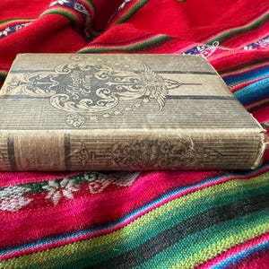 May include: A vintage hardcover book with a tan fabric cover featuring dark gray vertical stripes and intricate decorative scrollwork. The front cover displays the text Arlington Edition in a stylized font. The book spine shows ornate detailing.