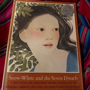 May include: A hardcover book titled "Snow-White and the Seven Dwarfs" with a portrait of a young woman on the cover. The illustration features the woman with long dark hair, rosy cheeks, and a blue dress. The book is a tale from the Brothers Grimm.