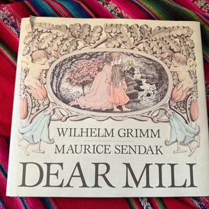 May include: A hardcover book titled "DEAR MILI" by Wilhelm Grimm and Maurice Sendak. The cover features an illustration of two figures in a forest scene, framed by an ornate border with cherubs. The book is set against a colorful textile background.