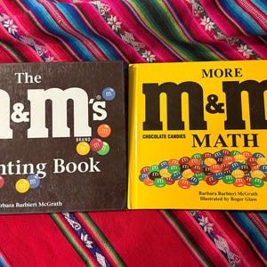 May include: Two children's books featuring the M&M's brand. One is a black book titled "The M&M's Counting Book" and the other is a yellow book titled "More M&M's Math". Both books have colorful M&M's candies on the cover.