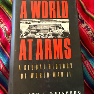 May include: A World at Arms book with a black cover and red lettering. The book's title is displayed in large red letters, with the subtitle "A Global History of World War II." The author's name is Gerhard L. Weinberg.