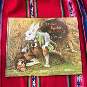 May include: A children's book titled "Doctor Rabbit's Foundling" by Jan Wahl, illustrated by Cyndy Szekerez. The cover features a white rabbit wearing a green vest and brown pants, holding a basket with a small animal inside.