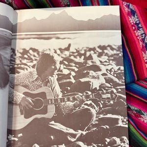 May include: Sepia-toned image of a person playing an acoustic guitar while seated on a rocky beach. The guitar is a light-colored wood with a dark pickguard. The background features a mountain range silhouette.