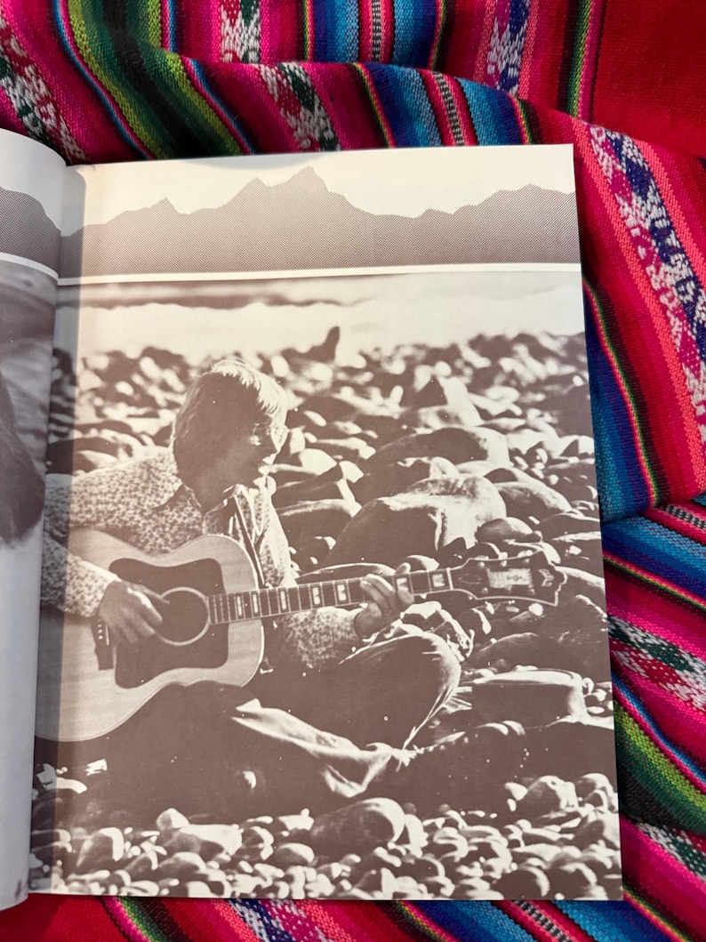 May include: Sepia-toned image of a person playing an acoustic guitar while seated on a rocky beach. The guitar is a light-colored wood with a dark pickguard. The background features a mountain range silhouette.