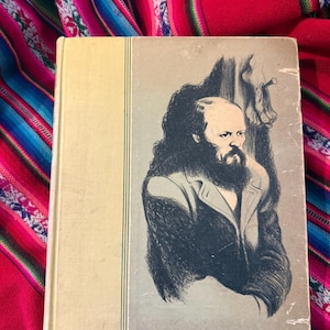 May include: A vintage book with a light yellow cover and a portrait of a bearded man on the front. The portrait is a black and white illustration. The book is resting on a colorful textile.