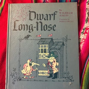 May include: A vintage book titled "Dwarf Long-Nose" by Wilhelm Hauff, translated by Doris Orgel. The cover is a muted blue with an illustration of a long-nosed figure and another character. The book has a decorative design and is set against a colorful textile background.