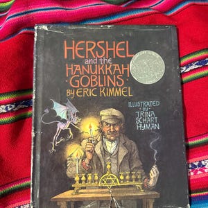 May include: A vintage hardcover book titled "Hershel and the Hanukkah Goblins" by Eric Kimmel, illustrated by Trina Schart Hyman. The cover features an illustration of a man lighting a menorah, with a goblin and text in orange and white.