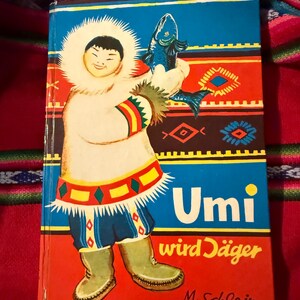 May include: Vintage children's book titled "Umi wird Jäger" featuring a colorful illustration of a person in Inuit clothing holding a blue fish. The cover has a red, blue, and white design with text in German.