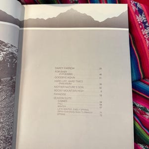 May include: Open book with a gray and white mountain range illustration at the top. The page features a list of song titles and page numbers, including "Darcy Farrow" and "Rocky Mountain High."