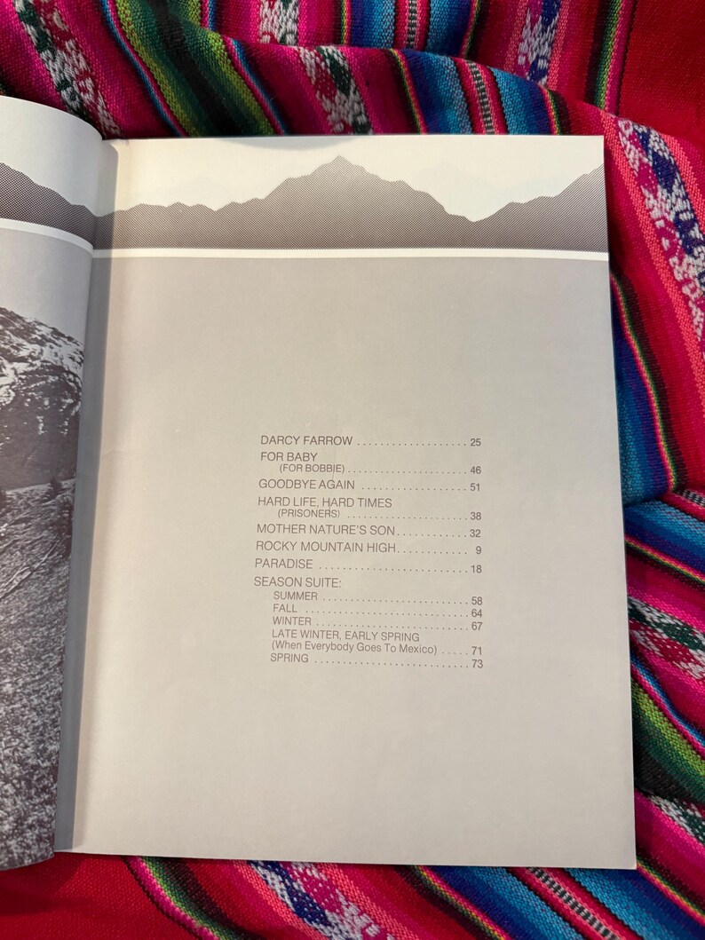 May include: Open book with a gray and white mountain range illustration at the top. The page features a list of song titles and page numbers, including "Darcy Farrow" and "Rocky Mountain High."