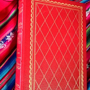 May include: A red leather-bound book with a gold-embossed design on the cover. The book is closed and lying on a colorful striped fabric.