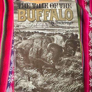 May include: A vintage book cover with a brown background featuring a black and white illustration of a buffalo. The title of the book is "The Time of the Buffalo" by Tom McHugh.
