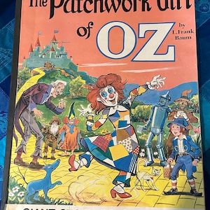May include: A vintage coloring book cover featuring a patchwork girl from the "The Patchwork Girl of Oz" story by L. Frank Baum. The cover features a colorful illustration of the patchwork girl, the Tin Woodman, the Scarecrow, and a blue dog. The book is titled "Giant Story Coloring Book".